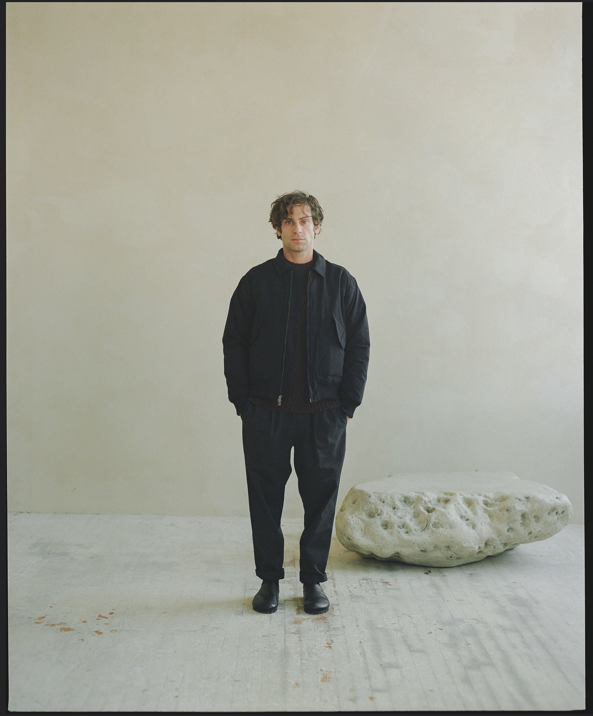 A man in a black bomber jacket and slouchy pants stands next to a rock.