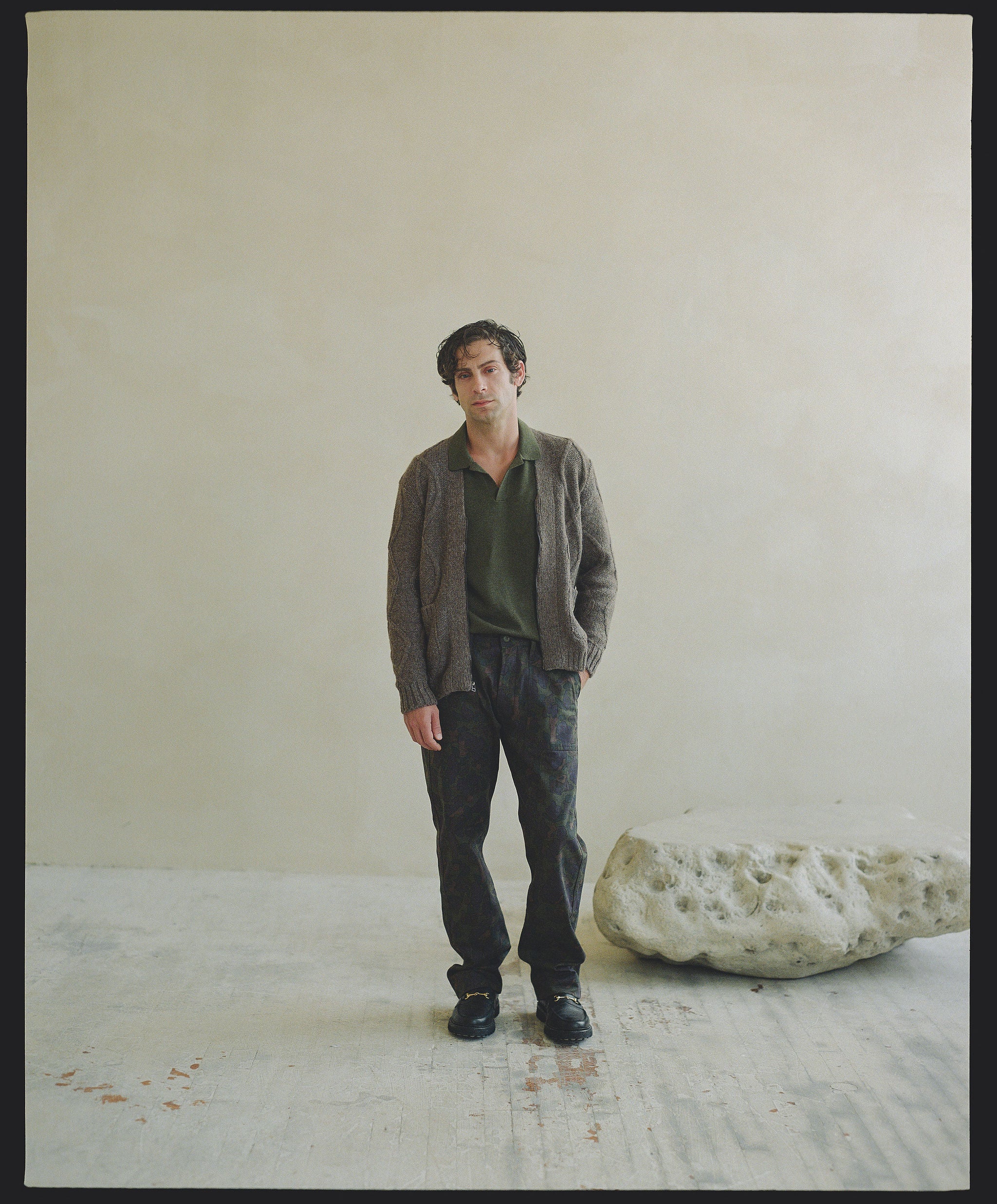 A man in an olive sweater and camouflage pants stands next to a rock.