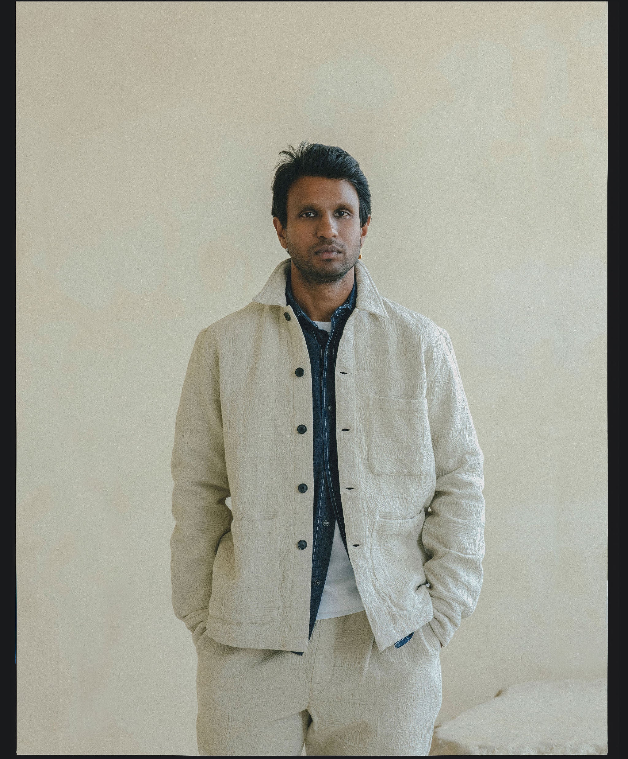 A man in a textured white jacket with a denim shirt underneath looks straight into the camera.