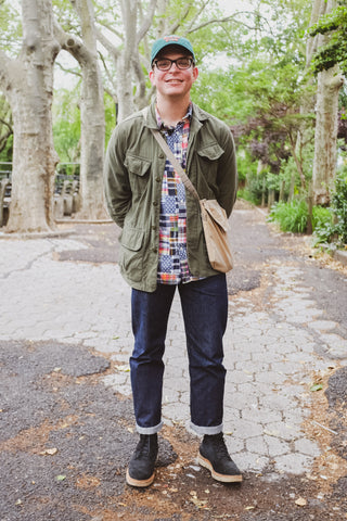 Chris stands and smiles in a park, showing off his personal outfit featuring a 3sixteen olive BDU jacket.