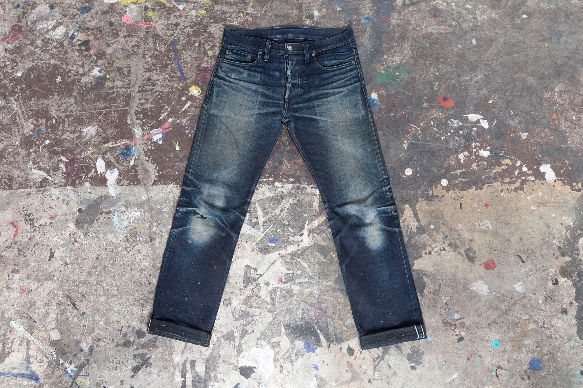Shot of a well-worn pair of SL-120x denim on a paint-splattered concrete background.