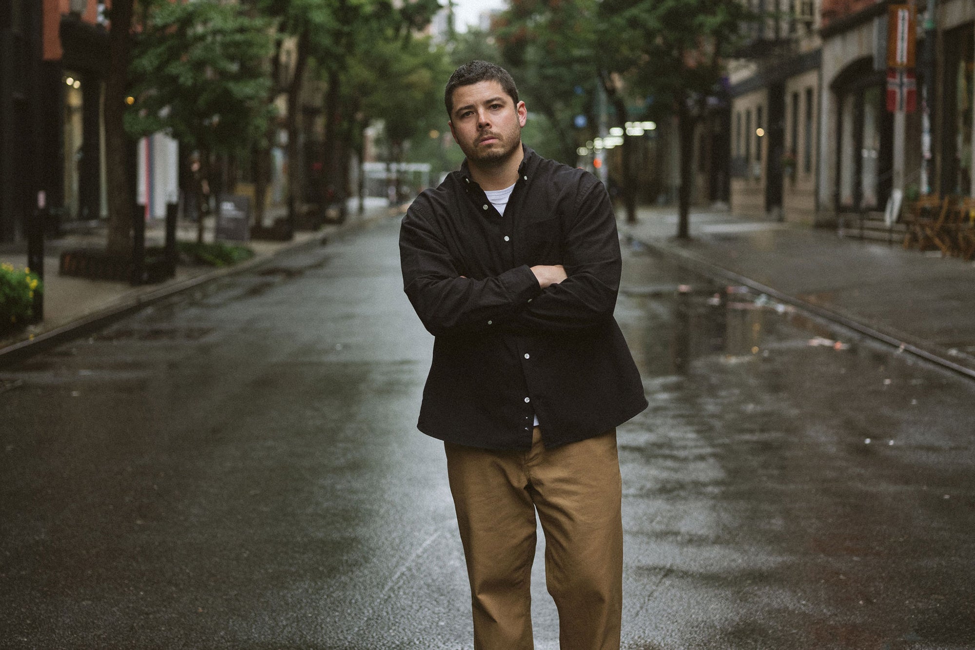 A man in a black oxford and tan pants stands on a rainy street.