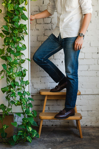 The man wears a white button down shirt, faded blue jeans, and oxblood tasseled loafers.