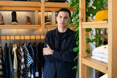 A handsome man in a navy shirt stands by a wooden rack of clothing.