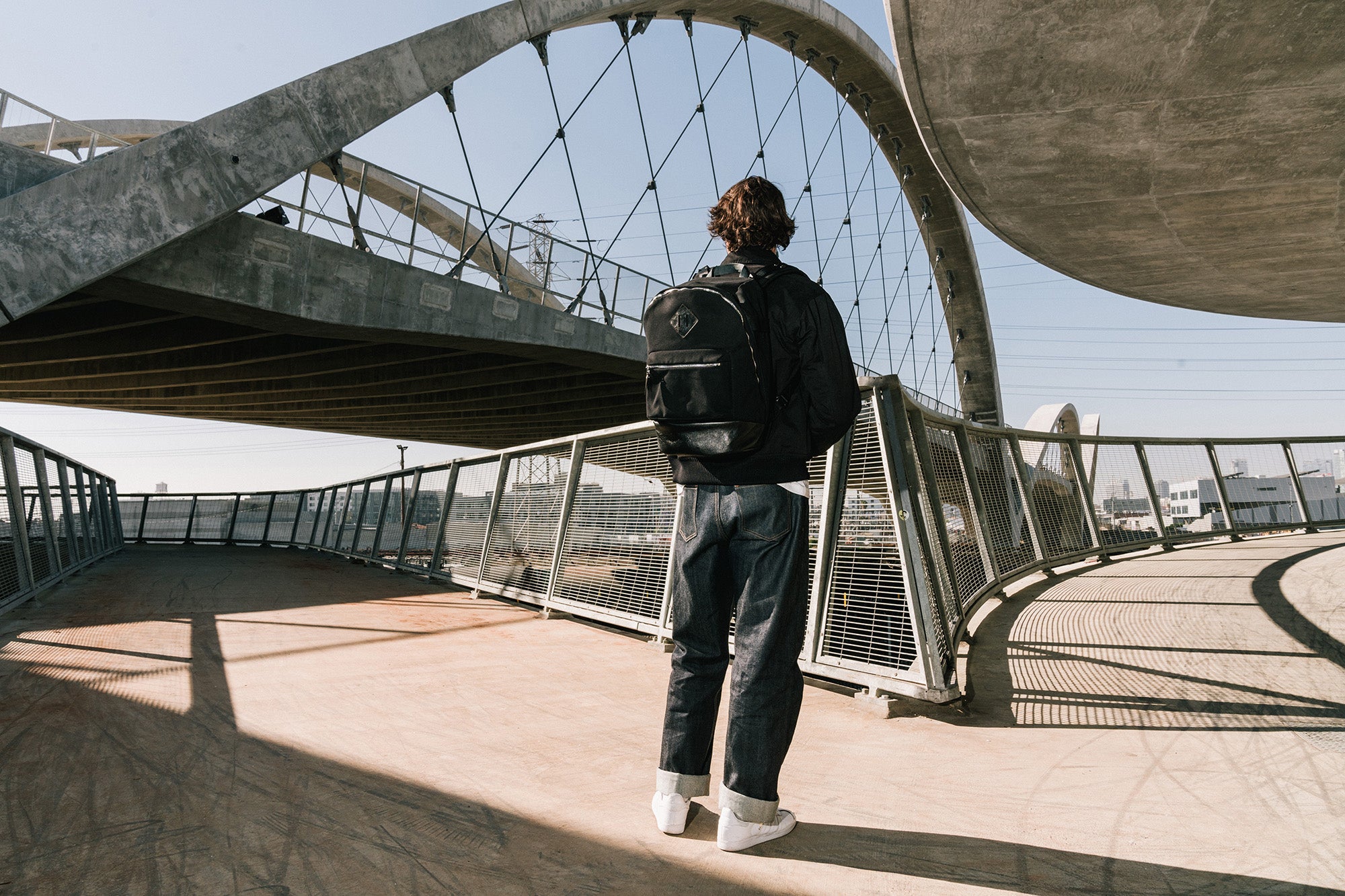 A man in a black jacket and a black backpack stands on a bridge.