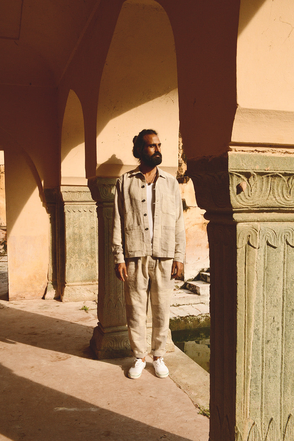 A man in a grey jacket and drawstring pants stands in a stone archway.