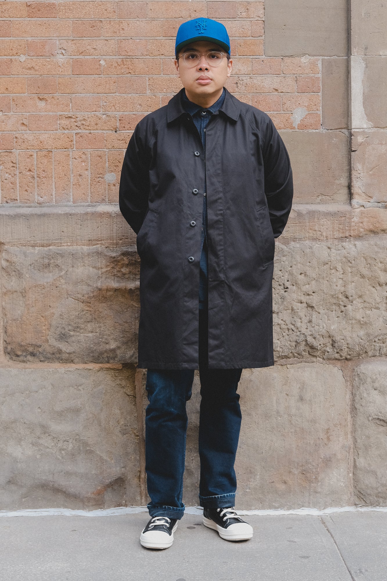 A man in a black mac coat and blue jeans stands against a brick wall.