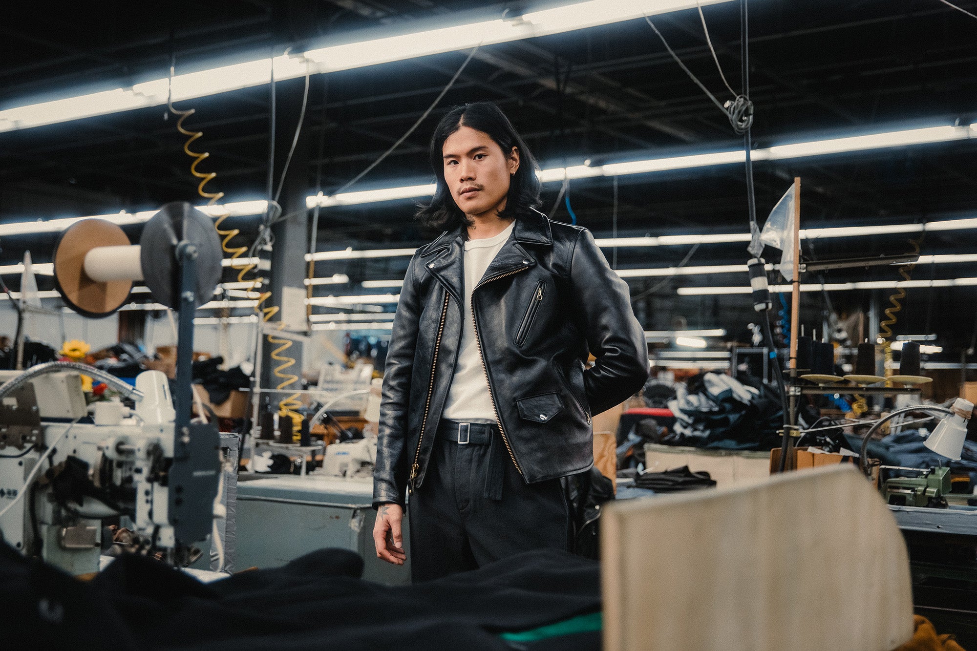 A handsome man in a black leather jacket stands on a factory floor.