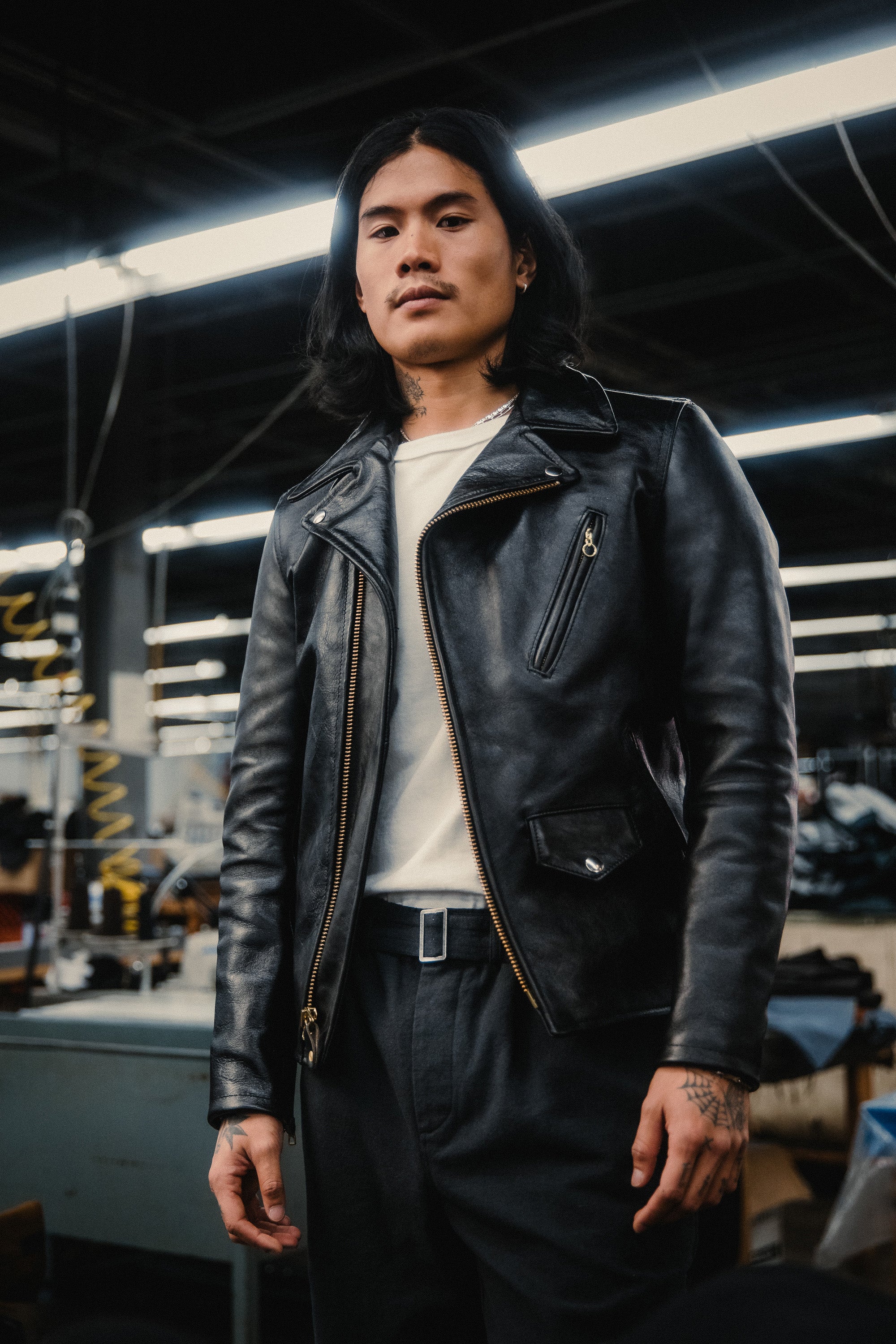 A man in a black leather jacket stands on a factory floor.