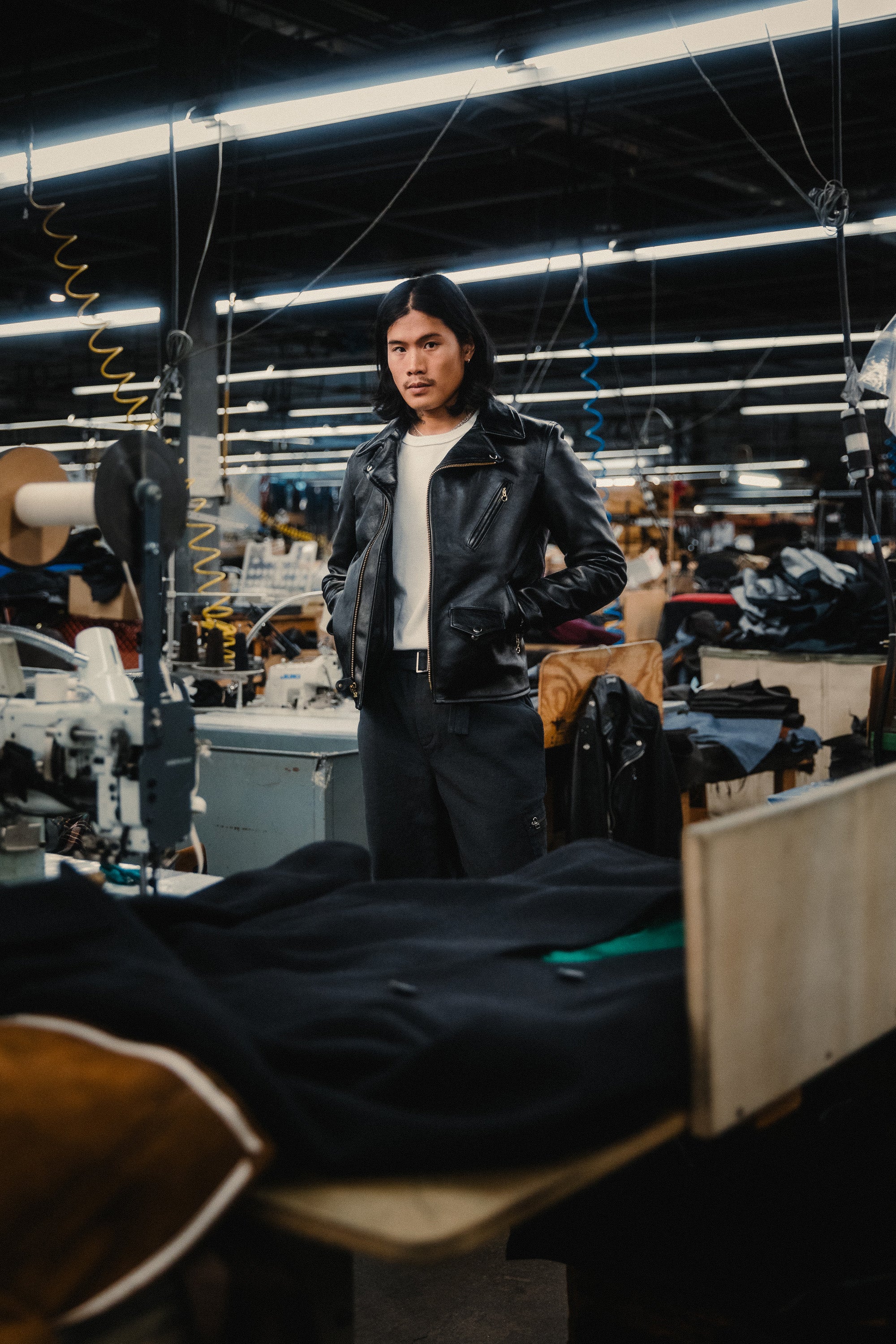 A man in a black leather jacket stands on a factory floor.