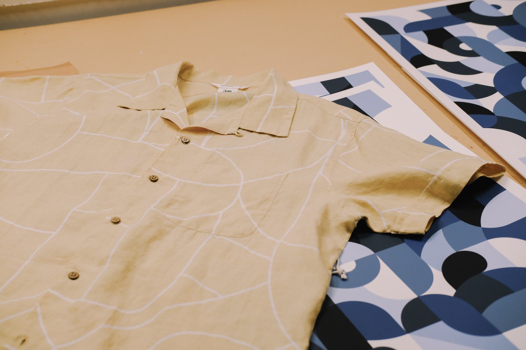 A beige printed shirt lays on a table over a series of blue art prints.