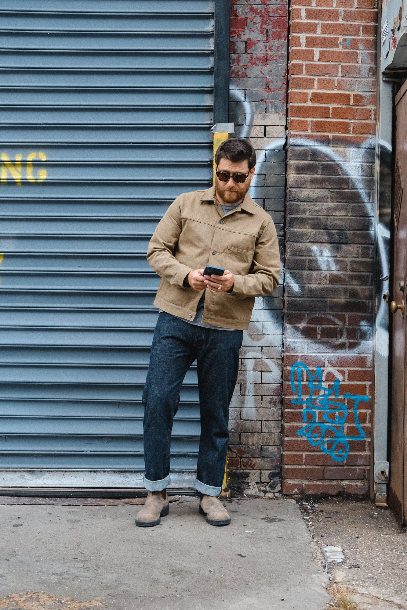 A man in a tan jacket and blue jeans stands outside a garage.