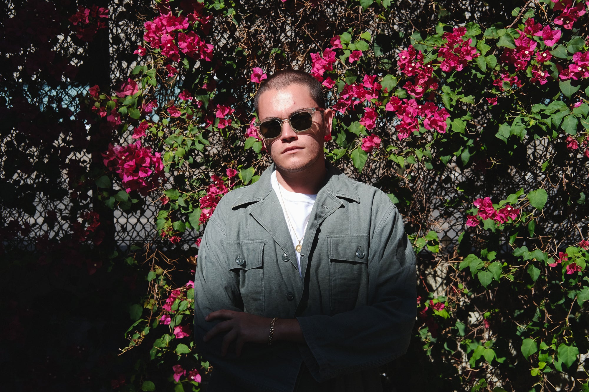 A man in sunglasses and an olive jacket stands in front of pink flowers.
