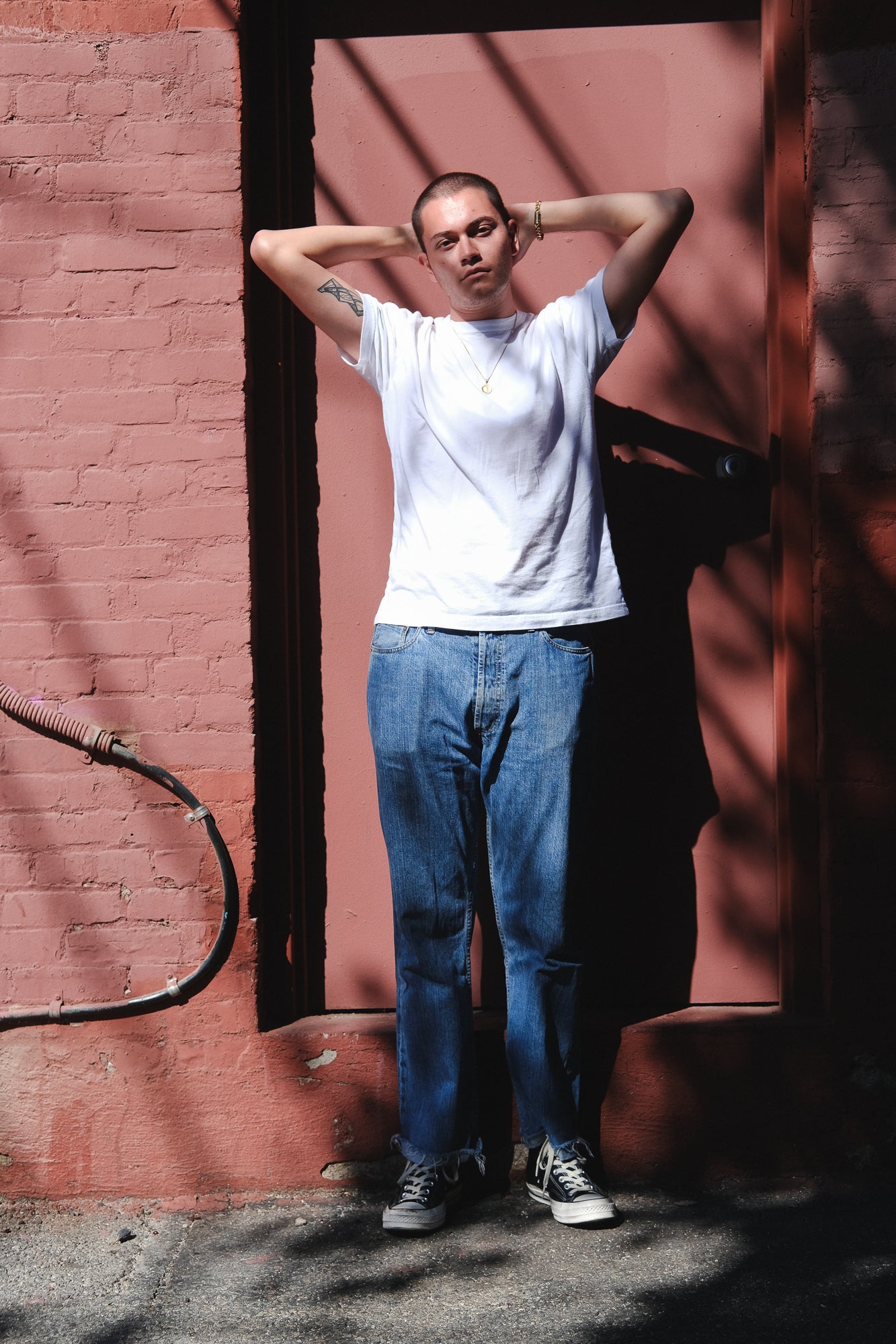 A man stands with hands behind his head against a red wall.