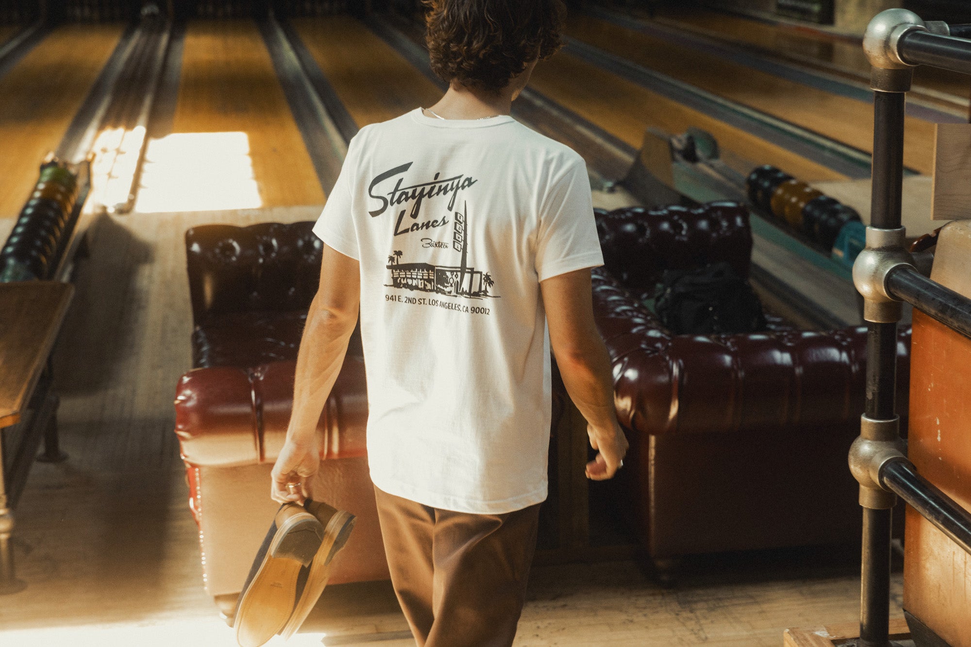 A man in a white graphic tee with "Stayinya Lanes" printed on the back walks down some stairs.