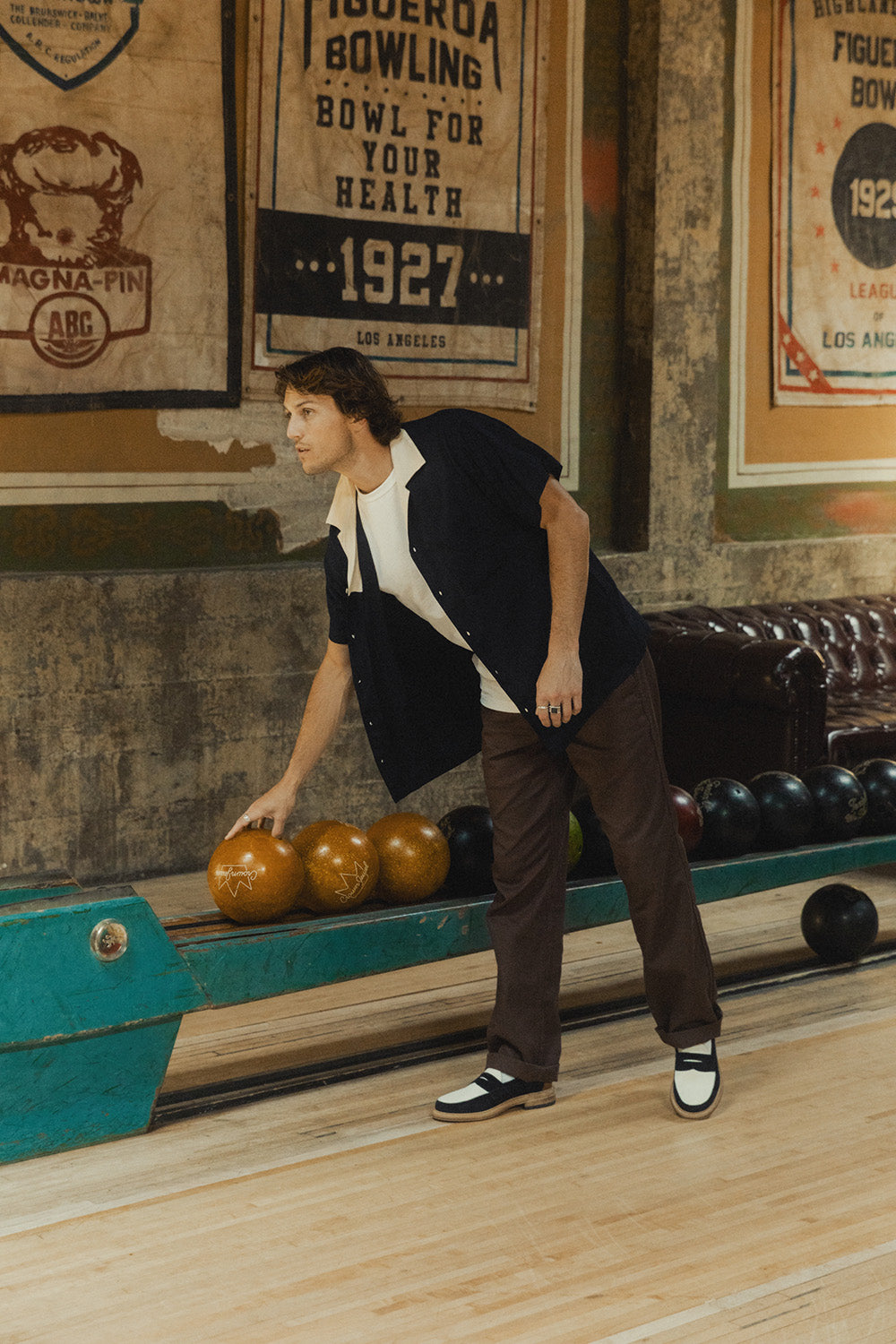 A man in a navy shirt and brown pants reaches for a bowling ball.