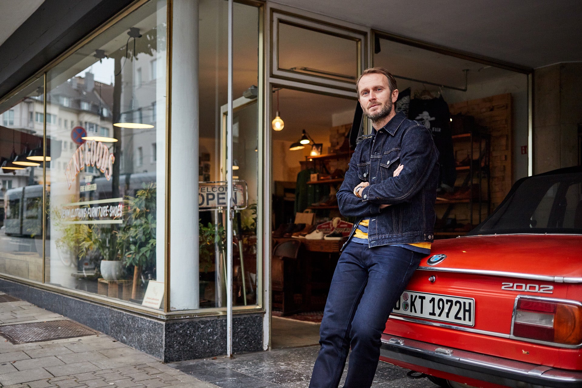 A man in a denim jacket and jeans stands with his vintage red BMW outside his shop.