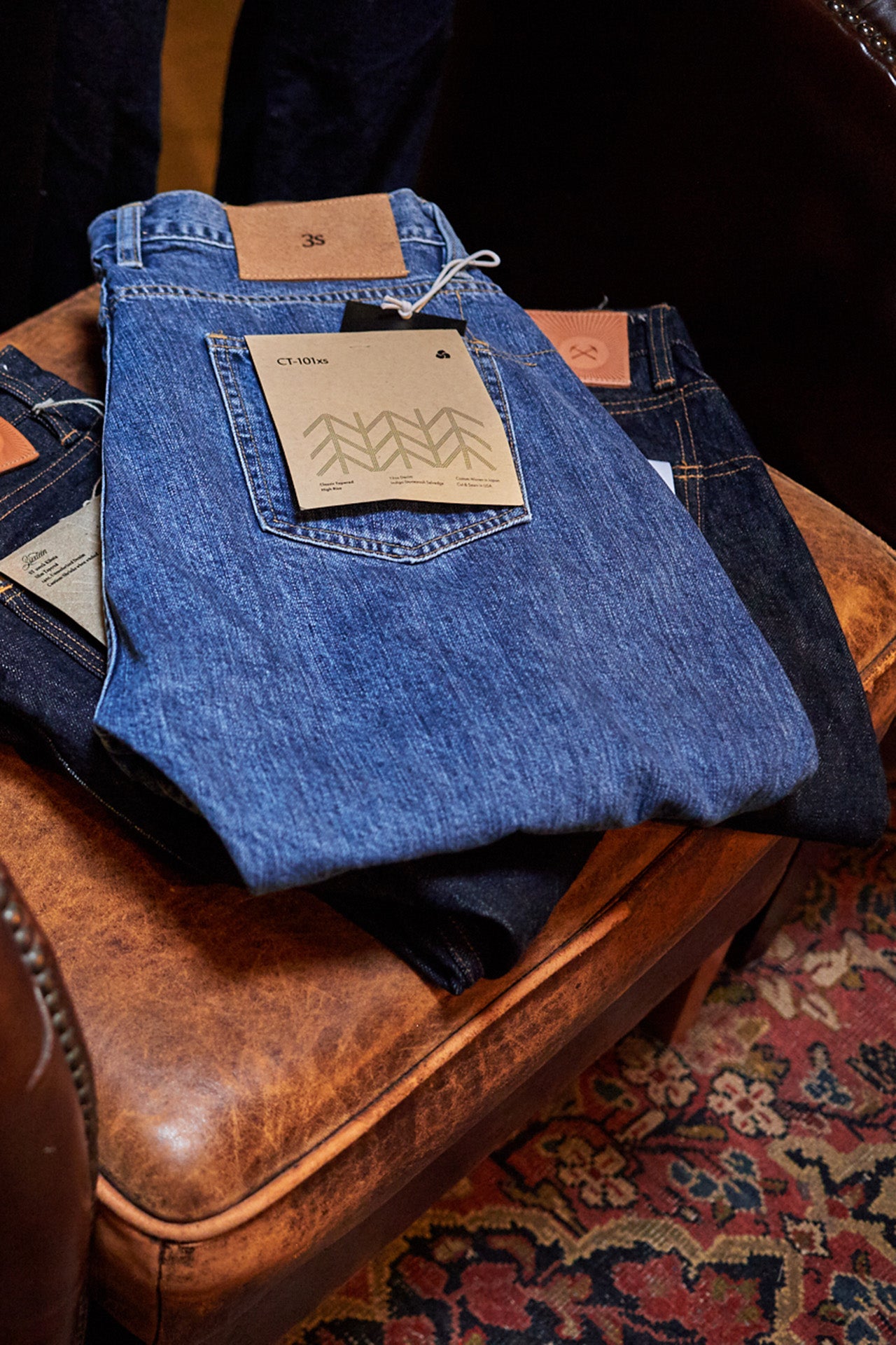 3sixteen blue jeans sit stacked on a beautiful aged leather ottoman.