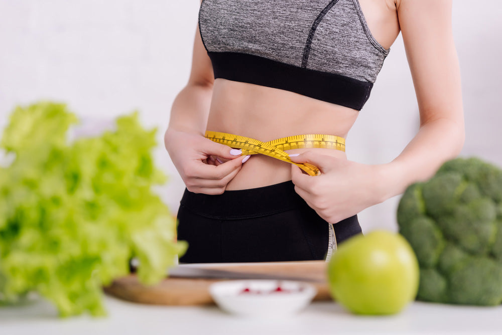 Image of a woman measuring her waist in front of a counter with healthy food