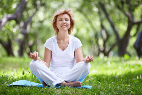 Image of a woman meditating in white on a sunny day on grass with trees behind her