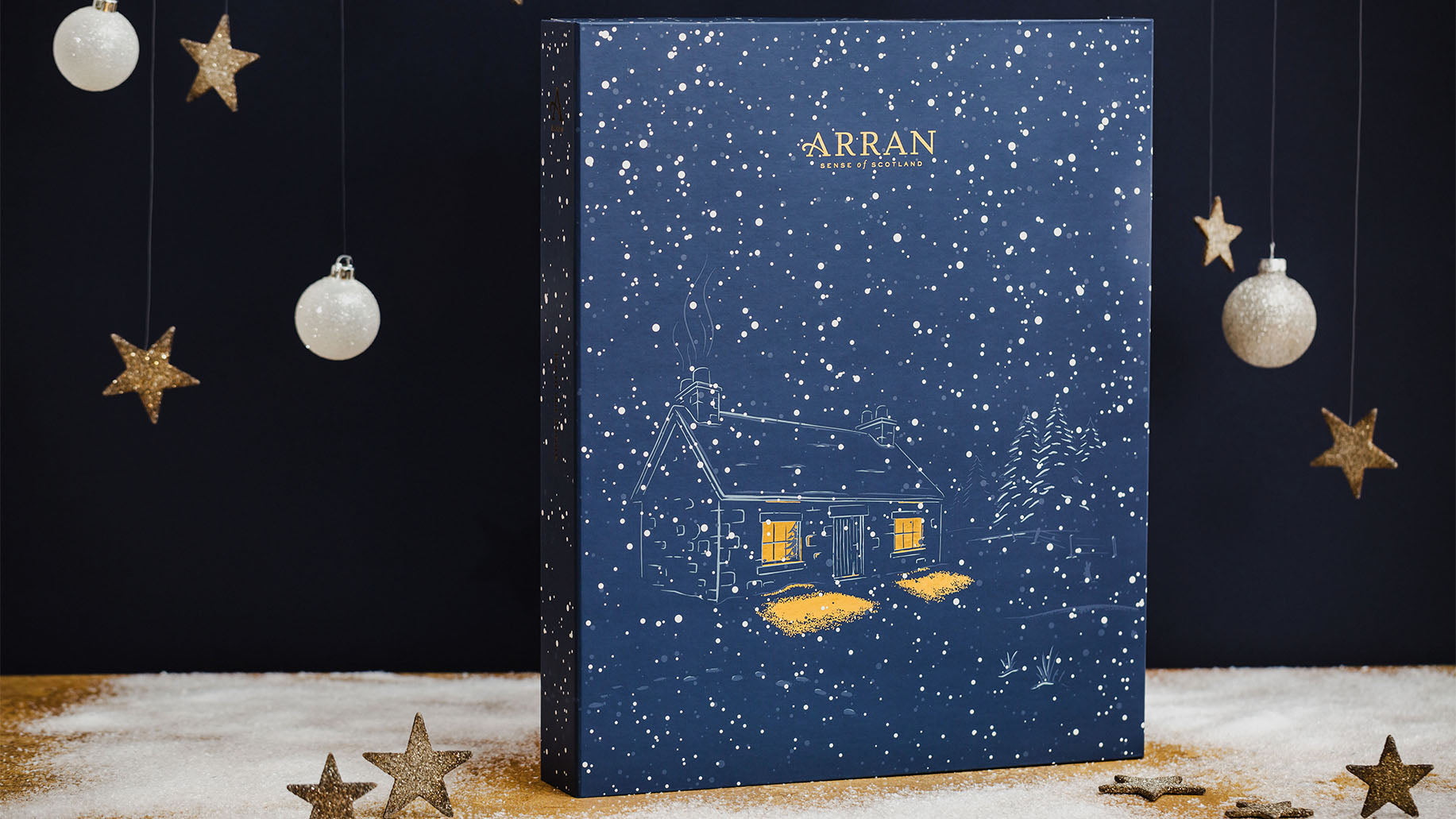 ARRAN Sense of Scotland 2022 Advent Calendar surrounded by snow, gold stars and white baubles