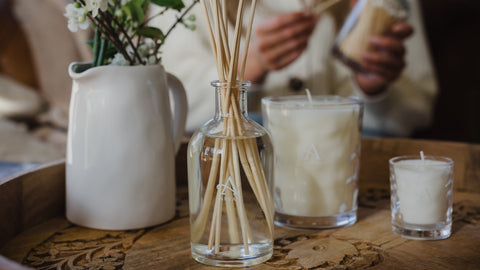 Image shows products within home fragrance collection on a round, wooden tray. From left to right, there is a rustic, ceramic milk jug with fresh white flowers, in the centre is a clear, glass diffuser with light wooden reeds coming out the top. To the right is a large 35cl cream coloured candle, in a dimpled, clear glass jar and next to this is the same candle but smaller (8cl). 