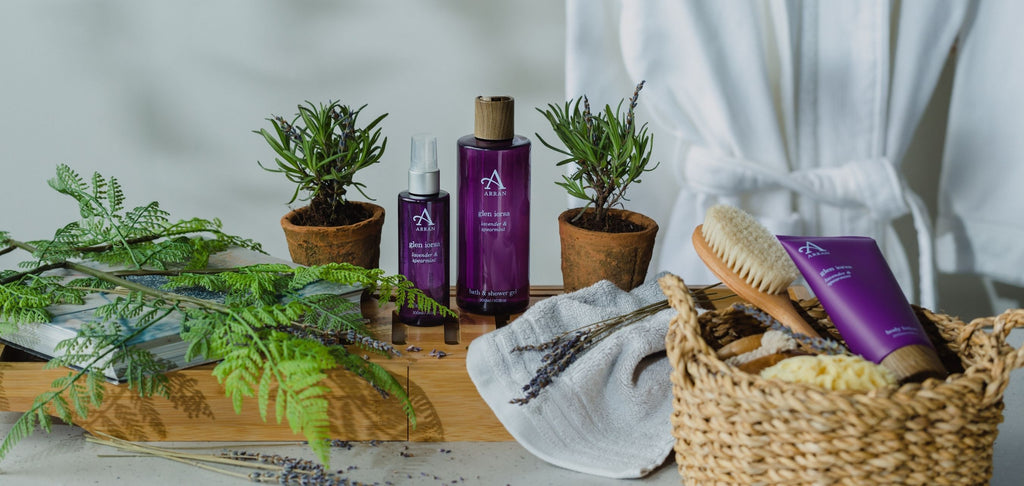 Image of lavender shower gel and body mist on wooden bath blank with white dressing robe in background.