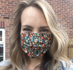 An image of a young woman with shoulder length dark blonde hair taken outside against a background of red bricks wearing a bright floral print cotton reusable washable face mask by Wiff Waff Designs.