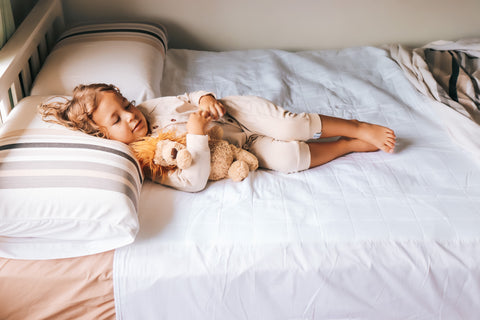 Toddler lying on their bed ontop of their white brolly sheet with wings holding their lion teddy