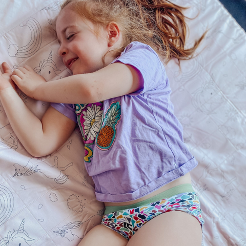 female toddler lying happily on top of a Brolly Sheet with Wings in Dusty Rose unicorn, with her Mosaic Night Training Pants