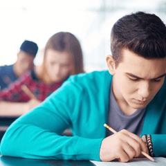 A male college student holding a pencil and sitting at a desk o take a test, with other students in the background
