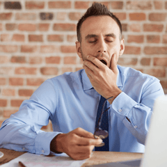 A melatonin-deficient businessman in a blue shirt and tie yawning at his desk due to excessive sleepiness. 