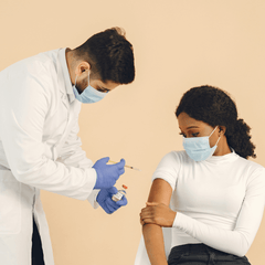 A brown-haired male doctor wearing a white lab coat, face mask and blue medical gloves stands and prepares to administer a COVID-19 vaccine to the arm of a brown-haired woman wearing a white shirt and face mask and sitting in a chair.