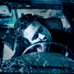 A sleeping man whose face is resting on the steering wheel of his car