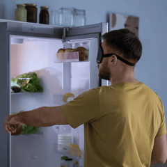 A sleepwalking man in a yellow shirt and sleep mask reaching into a kitchen fridge