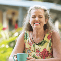 An older woman wearing a dress and calmly smiling as she relaxes over a cup of tea.