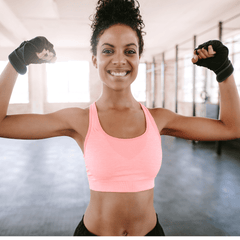 A smiling woman flexing her biceps in a gym, wearing a pink tank top and black fingerless gloves