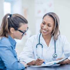 A smiling woman doctor in a white lab coat with a stethoscope, holding a pen and showing a file to a woman patient who is smiling and wearing glasses.