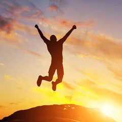 A silhouette of a person with their arms extended overhead  jumping off a large rock with the sunset and clouds in the background