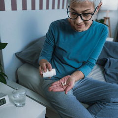 An elderly woman with gray hair, glasses, blue shirt and gray pants pouring melatonin pills from a bottle in one hand into her other hand as she sits in bed.