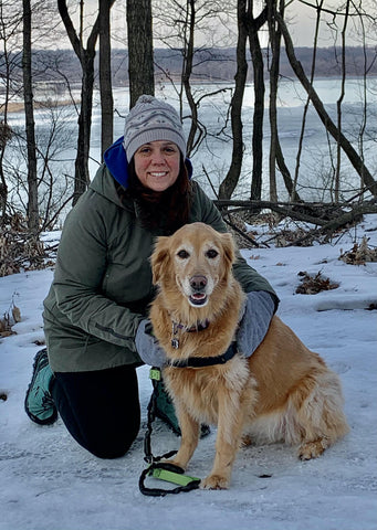 woman in gray hat and black pants kneeling next to Golden Retriever Bella in the snow