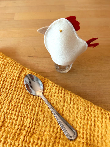 white felt chicken egg cozy shown covering an egg with a yellow napkin and spoon on a wooden table