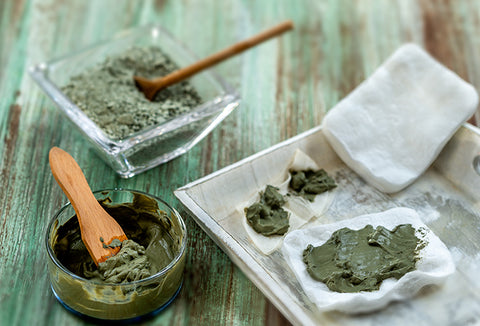 Two clear containers with wood spoons, mixing up green herbal poultices. A white tray and washcloths are nearby.