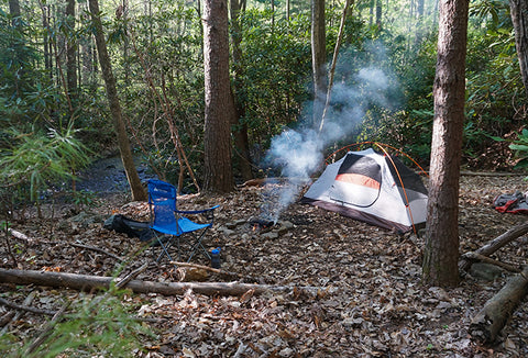 A wooded area with a tent, fire, and camp chair set up.