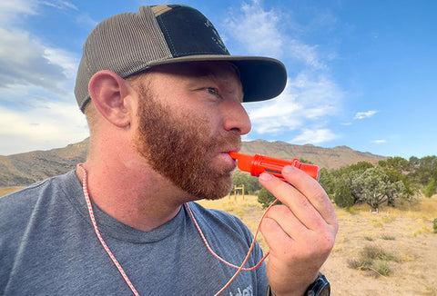 A bearded man with a baseball hat blowing an orange survival whistle in the desert.