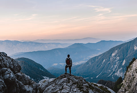 A man with a backpack standing on a mountain cliff, looking out at a mountainous sunset.