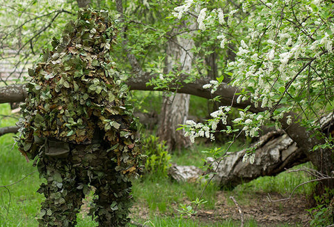 A person wearing a green, leafy ghillie suit to blend in to his green forest surroundings.