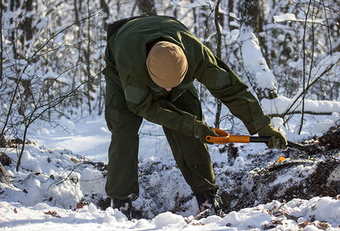A person digging a large hole in the snow in a wooded area.