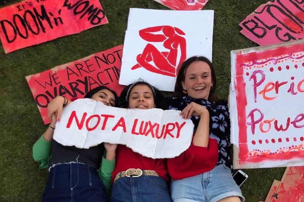 three girls holding a sign about periods saying not a luxury.