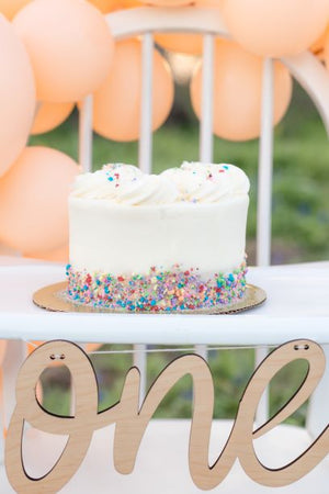 A white cake with small multicolored sprinkles on the bottom sits on the highchair with the wooden cursive one banner hanging.